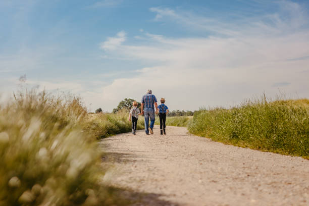 Grandfather with grandsons holding hands together while walking on dirt road beside field