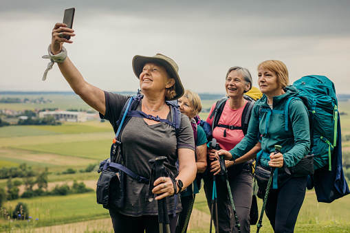 Mature female hikers making selfie photo together while standing on hill against clouds