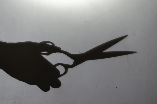 Blur, defocus, noise, grain effect. Dark silhouette of a man's hand with open scissors on a light gray wall. Man holding large scissors. Inside the room. Selective focus.
