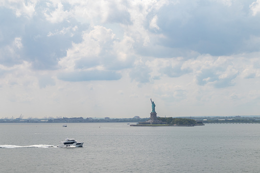 The statue of liberty in the distance along New York Harbor with boats and large clouds in the sky