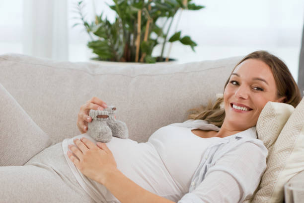istock Young, pregnant woman showing baby booties while lying down on the sofa 1354125189