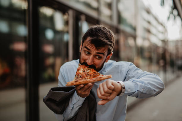 I have time for a quick meal Image of a young businessman in rush, eating a pizza slice and checking the time on his wristwatch. instant food stock pictures, royalty-free photos & images