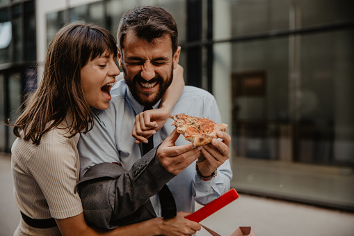 Beautiful brunette businesswoman with her arm around a handsome bearded colleague who is holding a pizza slice.