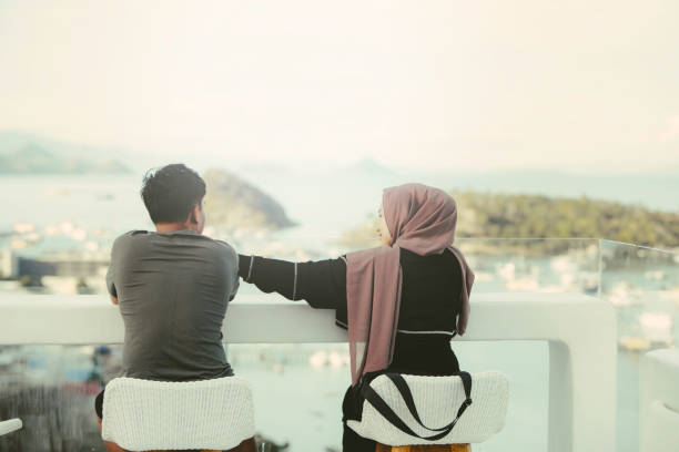 two young asian muslim couple sitting on the cafe  and looking the labuan bajo islands view on summer day two young asian muslim couple sitting on the cafe  and looking the labuan bajo islands view on summer day malay couple full body stock pictures, royalty-free photos & images