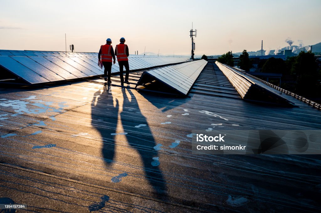 Male engineers walking along rows of photovoltaic panels Rear view of mature male engineers walking along the rows of photovoltaic panels on a rooftop of a solar plant. Solar Panel Stock Photo