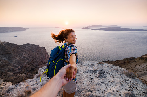 Happy female hiker holding hands with her unrecognizable boyfriend while being on a hill above the sea at sunset. Copy space.