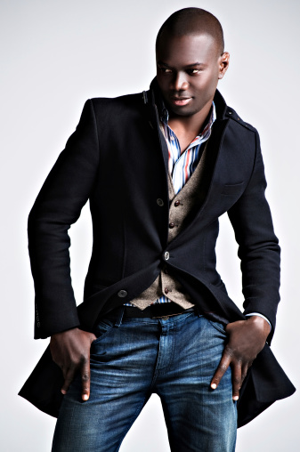 African male wearing winter fashion including a pair of jeans, vest, button down striped shirt, and a long black coat.