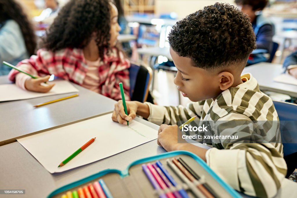 Focused Schoolboy Drawing Picture with Colored Pencils Close-up of 7 year old student in casual clothing sitting at child-size desk and using colored pencils to create a picture in class. Drawing - Activity Stock Photo