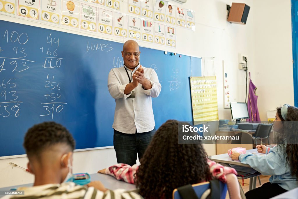 Smiling Teacher Applauding Young Student in Classroom Elementary age boys and girls sitting at child-size desks and learning arithmetic from encouraging instructor in late 40s standing at board. Teacher Stock Photo