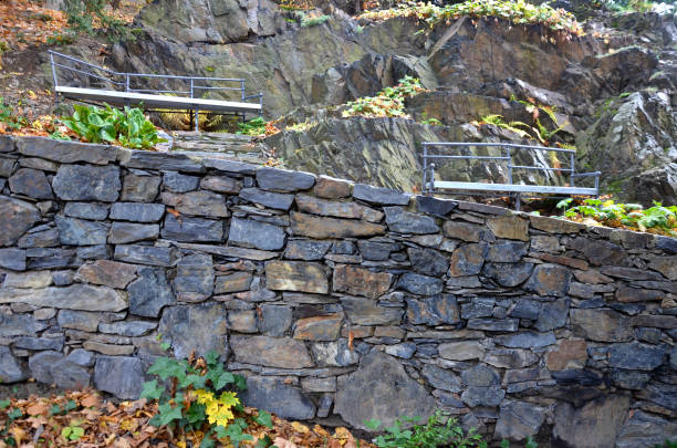 wooden benches of irregular geometric polygons on stone gray pavement in a park with low retaining walls stacked of porphyry. ornamental gardens with stairs and natural rocks of a wild character wooden benches of irregular geometric polygons on stone gray pavement in a park with low retaining walls stacked of porphyry. ornamental gardens with stairs and natural rocks of a wild character, bergenia cordifolia, dryopteris filix-mas, polygonal retaining walls stock pictures, royalty-free photos & images