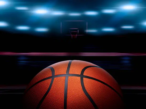 An indoor basketball court with an orange ball on an unmarked wooden floor under illuminated floodlights