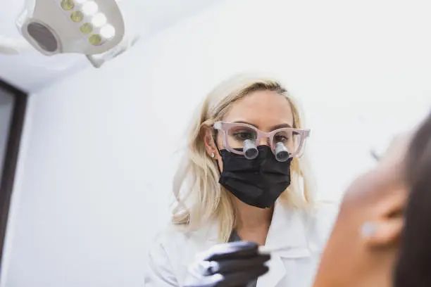 A dental exam from the female patient's point-of-view.  The mid adult female dentist wears a protective mask and loupes as she prepares to use her angled mirror.