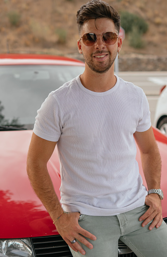 An attractive guy with sunglasses smiling sitting in the front of his car while looking at the camera in a viewpoint.