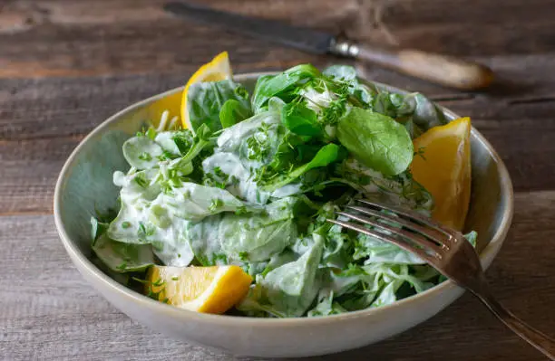 Fresh and homemade salad bowl with green lettuce, herbs and yogurt dressing. Served with lemons isolated on wooden table. Closeup view.