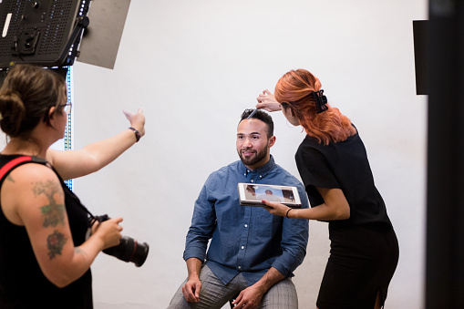 The mid adult female assistant holds a digital tablet as she follows the mid adult female photographer's instructions.  They are preparing the mid adult man for a promotional photo shoot.