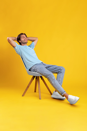 Vertical Portrait Of Happy Casual Millennial Man Relaxing Sitting In Chair Holding Hands Behind Head Leaning Back, Posing With Eyes Closed On Yellow Orange Studio Wall Background, Enjoying Silence