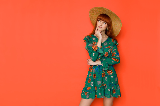 Young red haired woman in green summer dress and straw hat is thinking and looking to the side. Three quarter length studio shot on orange background.