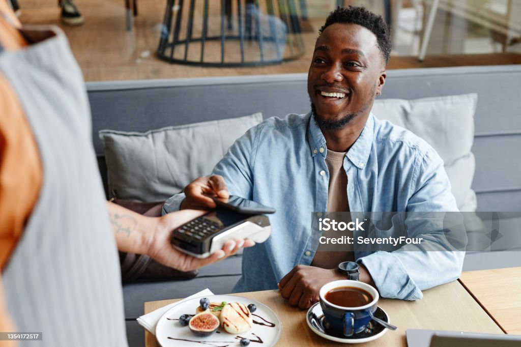 Man Paying For Order In Cafe Joyful young Black man sitting at table in cozy cafe paying for lunch using his smartphone Paying Stock Photo