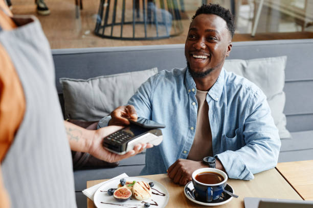 Man Paying For Order In Cafe Joyful young Black man sitting at table in cozy cafe paying for lunch using his smartphone paying stock pictures, royalty-free photos & images