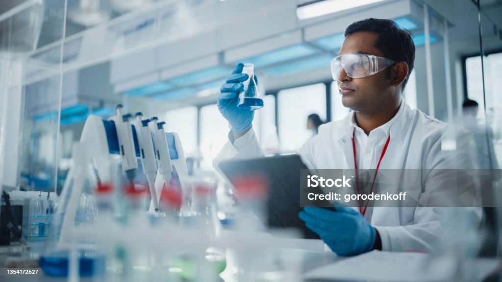 Medical Research Laboratory: Portrait of a Handsome Male Scientist Using Digital Tablet Computer, Analysing Liquid Biochemicals in a Laboratory Flask. Advanced Scientific Biotechnology Laboratory. Scientist Stock Photo