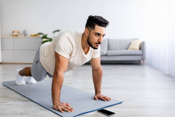 istock Strength workout. Fit young Arab man doing push ups or plank at home, following video tutorial on smartphone, free space 1354172878