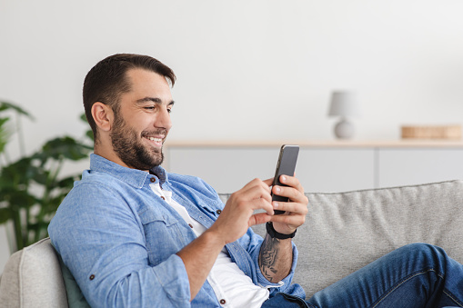 Glad mature adult caucasian man with beard sitting on sofa, chatting on smartphone in living room interior, profile. Social distance, video call, social networks, gadgets and new app at free time