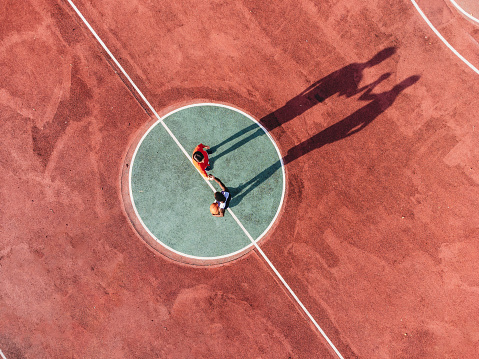 Two friends are playing basketball together, holding hands before the start - Aerial point of view. They are standing in the center of the basketball field.