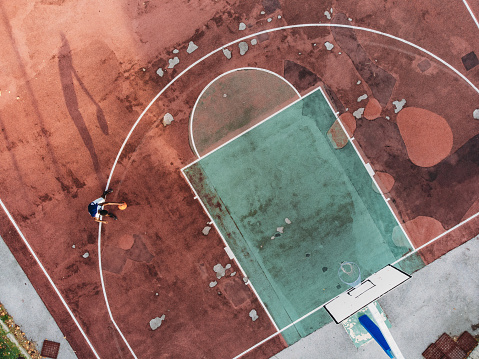 A young adult man is playing basketball in a pick-up basketball court.  Aerial point of view.