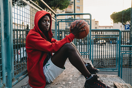 Portrait of a young adult man while he's sitting on the basketball court's bleachers. He's wearing a red hooded sweatshirt. He's looking at camera.