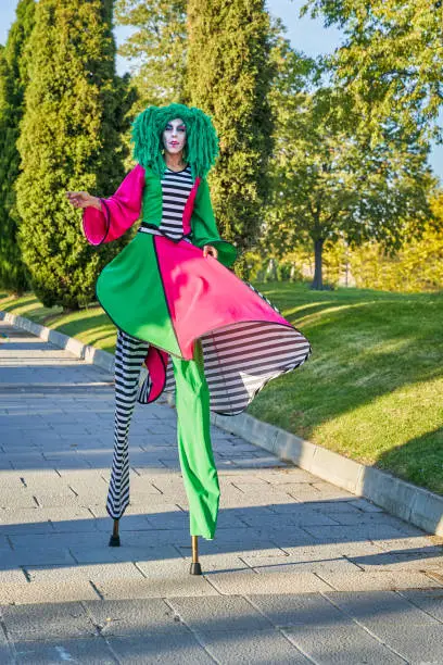 Full length of female clown with painted face in funny costume and green wig, walking on stilts on paved path in park on sunny day