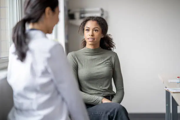 A young female patient sits casually with her doctor as they discuss her mental health.  She is seated in a chair in front of her doctor as they talk about her needs.  The doctor is wearing a white lab coat and has her back to the camera as the two discuss possible plans of care to navigate the woman's struggles.