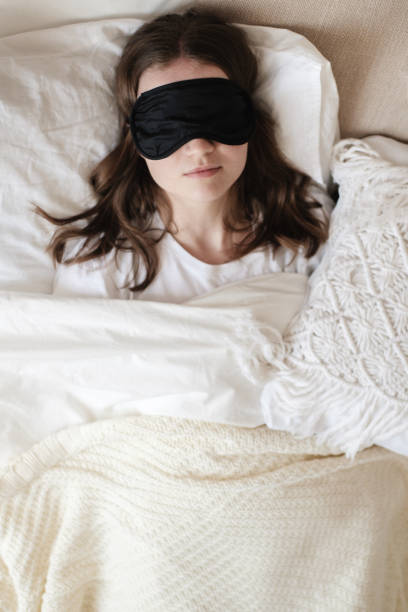 Young beautiful girl lying in comfortable bed wearing sleep mask in the morning at cozy room. Concept of rest at home, sleep, insomnia, self-isolation, stay home during quarantine, pandemic. stock photo