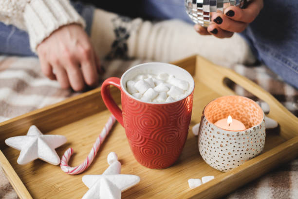 Young woman sits on plaid in cozy knitted woolen white sweater and holds cup of cocoa with marshmallows. Hygge banner New Year, cozy preparation for holidays. Candles, Christmas balls in wooden tray. stock photo