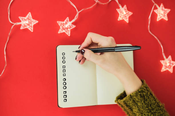 Female hands with manicure in green sweater write in notebook wish list, goals or promises for New Year on red background and garland in form of stars. Concept of to-do list. New life from new year. stock photo