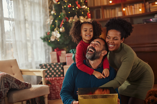 Mixed race family during Christmastime. Wife and daughter hugging smiling man who gets gift from them
