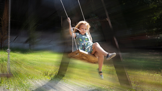Beautiful little girl in flight on a swing against the background of a blurred in motion park with large trees in the sun's rays. Joyful, carefree, happy childhood.