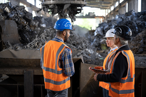 Male shredder manager talking with supervisor and female safety coordinator while scrap magnet carrying metal in background at the recycling facility.