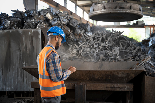 Side view of male shredder manager using digital tablet while supervising the industrial magnet taking scrap metal at the recycling facility