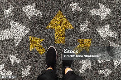 istock Female foots in black boots standing on road with arrow markings pointing in different directions with three options for arrow direction, left, right or move forward, decision making 1354189797