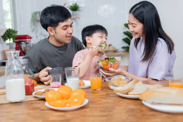 Asian Thai family parents take care young boy kid like to eat vegetable breakfast and drink milk in morning stock photo