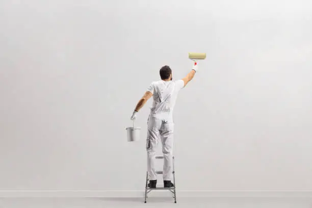 Photo of Rear view shot of a painter holding a bucket and painting a wall on a leader