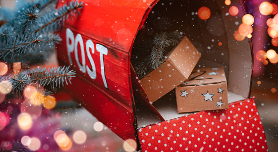red Christmas mailbox with gift boxes and a greeting card from Santa Claus. Christmas lights bokeh. Holiday delivery. Close-up.