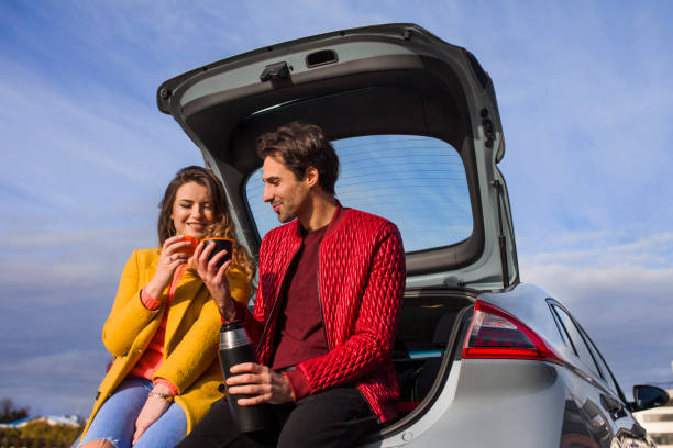 Young couple sitting on a car trunk, drinking tea from a thermos cups