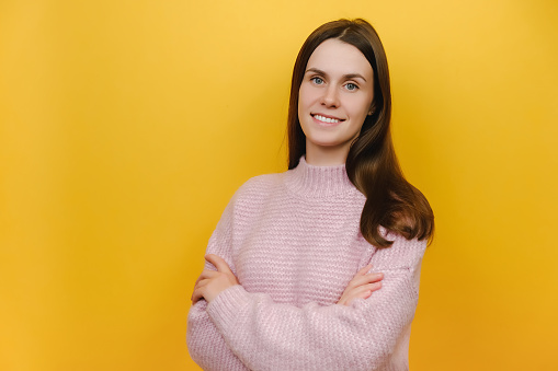 Side view smiling happy cheerful young caucasian woman wearing pink knitted sweater hold hands crossed folded, posing isolated over yellow color background wall in studio. People lifestyle concept