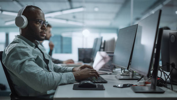 istock Office: Energetic Black IT Programmer Puts on Headphones Starts Working on Desktop Computer. Male Website Developer, Software Engineer Developing App, Video Game. Listening to Podcast, Music 1354205012