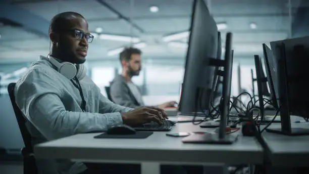 Photo of Diverse Office: Black IT Programmer Working on Desktop Computer. Male Specialist Creating Innovative Software Engineer Developing App, Program, Video Game. Terminal with Coding Language.