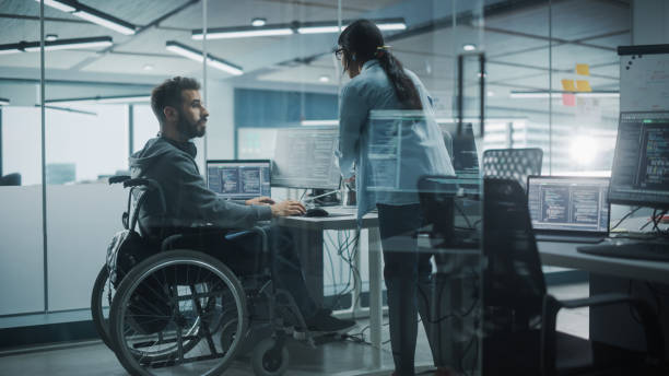 Disability-Friendly Office: Happy IT Programmer with Disability in a Wheelchair Talks with Colleague while Working on Computer. Software Engineers Develop Innovative App, Program. oficina amigable para la discapacidad: feliz programador de ti con discapacidad en una silla de ruedas habla con un colega mientras trabaja en la computadora. los ingenieros de software desarrollan una aplicación innovadora, un programa.