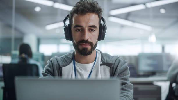 istock Office: Portrait of Smiling IT Programmer Wearing Headphones Working on Desktop Computer. Male Website Developer, Software Engineer Developing App, Video Game. Listening to Podcast, Music. Close-up 1354205486