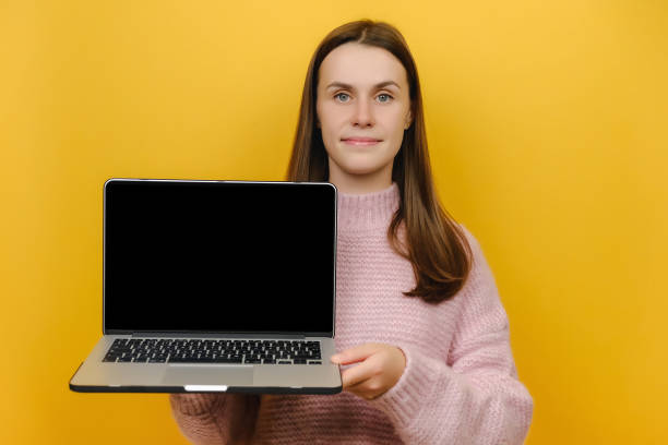 istock Smiling cute young female in pink knitted sweater hold laptop pc computer with blank empty screen, posing isolated over yellow color background studio. People lifestyle concept. Mock up copy space 1354206537