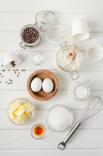 istock Ingredients for making muffins with chocolate chips. Eggs, butter, sugar, flour, vanilla, chocolate chips on a white wooden background. Recipe step by step. Top view. 1354206828